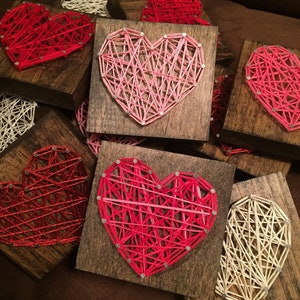 May include: A collection of small wooden squares with string art hearts in various colors, including pink, red, and white. The hearts are created by wrapping string around nails hammered into the wood.