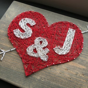 May include: A handmade string art heart with red thread, featuring the letters "S & J" in white thread. An arrow pierces the heart, all mounted on a wooden board. A unique, personalized piece.
