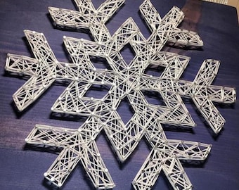 Peut inclure: Flocon de neige en fil blanc sur fond bleu. Le flocon de neige est fait de fil blanc enroulé autour de clous enfoncés dans le bois. Le texte "STRINGS BY SAMANTHA" est dans le coin supérieur droit de l'image.