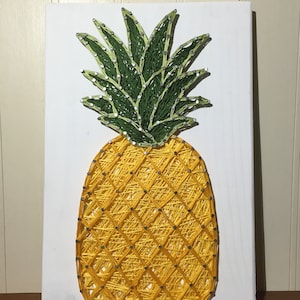 Op de afbeelding: Een gele en groene string art ananas op een witte houten achtergrond. De ananas is gemaakt met spijkers en touw, en de bladeren zijn groen en geel.