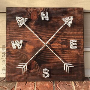 May include: A wooden sign with a compass design created with string art. The sign features the letters N, S, E, and W, each with an arrow pointing in the corresponding direction.