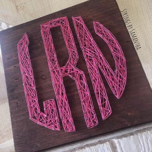 May include: A wooden sign featuring the letters "LRN" made from pink string. The letters are attached to a dark brown stained wooden board. The sign is square and the letters are in a sans-serif font.