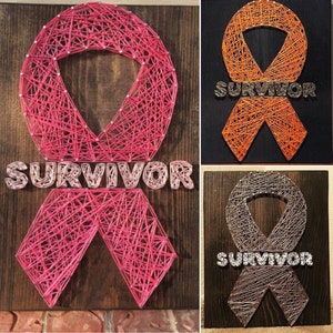 以下が含まれることがあります： ピンク、オレンジ、グレーのリボンを表現した3つの糸アート作品。各リボン下に「SURVIVOR」の文字が書かれている。