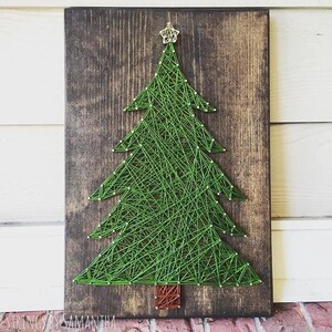Op de afbeelding: Een handgemaakt kerstboom string art stuk. De groene draadboom is gemonteerd op een donker gebeitste houten plank. De boom is bekroond met een gouden ster en heeft witte stip accenten. De basis van de boom is bruin.