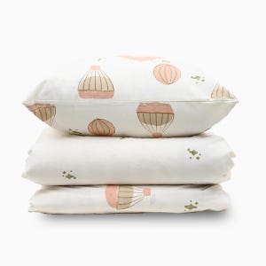 May include: A stack of white bedding featuring a repeating pattern of light pink and tan hot air balloons interspersed with small green star shapes. The set includes a pillowcase on top and folded linens underneath, all displaying the same print.