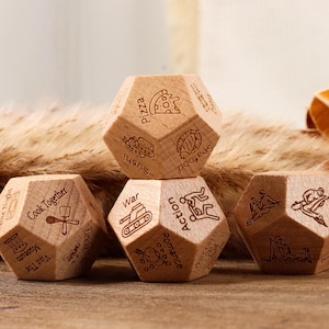 May include: Wooden, multi-sided dice with engraved icons and text. The dice feature images of pizza, sushi, and a tank, among others. The dice are arranged on a wooden surface with dried plants in the background.