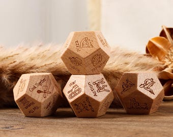 May include: Wooden, multi-sided dice with engraved icons and text. The dice feature images of pizza, sushi, and a tank, among others. The dice are arranged on a wooden surface with dried plants in the background.