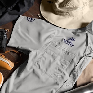 May include: A gray short-sleeved button-down shirt with a pocket on the left side. The shirt has a navy blue embroidered logo that says "Big Fish Brewing Co."