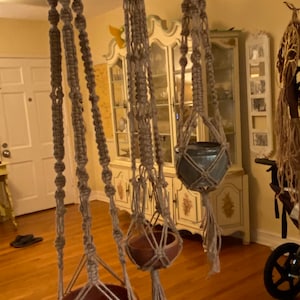 May include: Three macrame plant hangers with wooden pots hanging from the ceiling. The hangers are made of natural twine and have a simple, bohemian style.