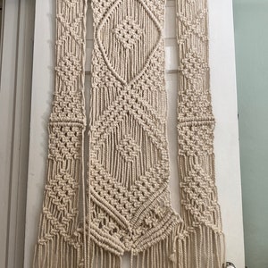 May include: A beige macrame wall hanging with a geometric diamond pattern. The hanging is made of natural cotton rope and has long fringe tassels.