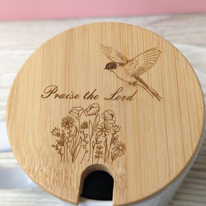 Pu&ograve; includere: Un coperchio per tazza in legno con un design di uccello e fiori. Il coperchio ha un'apertura per una cannuccia e il testo "Praise the Lord" &egrave; inciso sul coperchio.