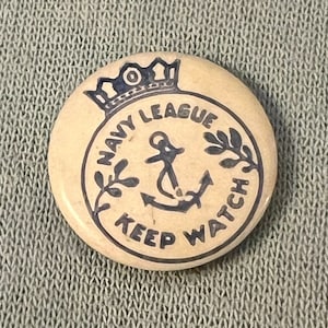 May include: A circular, cream-coloured Navy League button with dark blue text and illustrations. The button features a crown, an anchor, and the words "NAVY LEAGUE" and "KEEP WATCH" in a circular design.