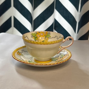 May include: A vintage teacup and saucer set. The cream-coloured cup and saucer feature yellow floral and trim details. The cup has a curved handle with red accents. The saucer has a matching floral design.