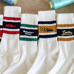 Op de afbeelding: Crèmekleurige sokken met gestreepte banden in rood, zwart, groen en oranje. Sommige sokken hebben geborduurde tekst: "DAD", "MUM", "Embroidery custom", "Maria", "Emilia" en "RB". De sokken worden getoond met garenklosjes en bloemen.