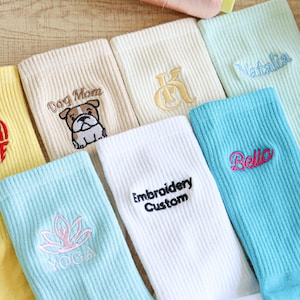 Könnte beinhalten: Eine Sammlung bunter Socken mit gestickten Designs und Text. Die Socken sind in Gelb-, Beige-, Weiß- und Blautönen gehalten. Einige Socken zeigen die Wörter "HURRICANE", "Dog Mom", "Embroidery Custom", "Bella" und "YOGA".