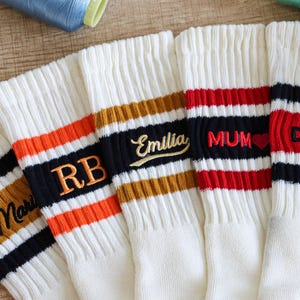 Könnte beinhalten: Cremefarbene Sportsocken mit gestickten Namen und Initialen in verschiedenen Farben. Die Socken haben gestreifte Bänder in Orange, Schwarz und Rot. Die Namen "Marl", "RB", "Emilia", "MUM" und "DAD" sind auf die Socken gestickt.