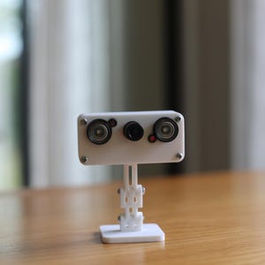 May include: A white rectangular camera module featuring two circular lenses on the sides and a smaller central lens, mounted on an adjustable white articulated stand with a square base. The device is secured with visible silver metal screws.