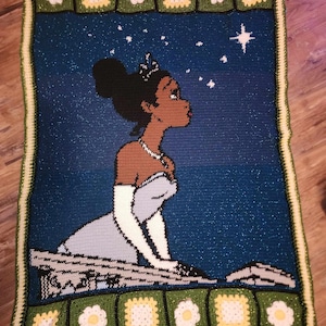 May include: A crocheted blanket featuring a cartoon image of a princess with dark skin, a blue dress, and a crown. The blanket has a green border with white flowers.