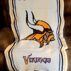 May include: White crocheted blanket with a purple and gold Minnesota Vikings logo and the words "Minnesota Vikings". The blanket has a black, white, purple, and gold border.