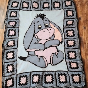 May include: A blue and pink crocheted blanket featuring a cartoon image of Eeyore the donkey from Winnie the Pooh. The blanket is made with a square pattern and has a black border.