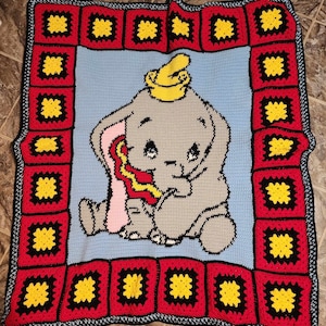 Puede incluir: Una manta de bebé de ganchillo con un Dumbo el elefante gris y rosa con un sombrero amarillo. La manta tiene un borde rojo y negro con cuadrados amarillos.