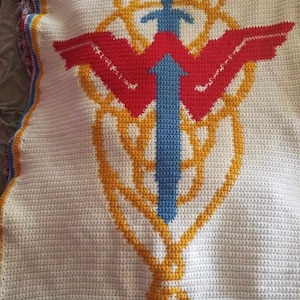 Pode incluir: Uma manta de crochê branca com um design amarelo, vermelho e azul. O design apresenta uma espada com asas e um círculo ao redor.