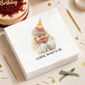 May include: A stack of white paper napkins with a printed image of a smiling child wearing a party hat with the number 30. The text "LOOK WHO'S 30" is printed below the image. Gold candles, confetti, and a cake are in the background.