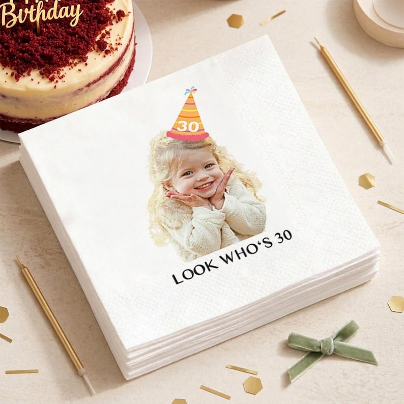 May include: A stack of white paper napkins with a printed image of a smiling child wearing a party hat with the number 30. The text "LOOK WHO'S 30" is printed below the image. Gold candles, confetti, and a cake are in the background.