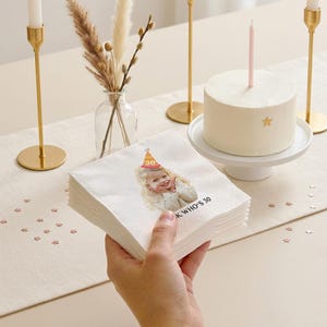 May include: A stack of white paper napkins with a printed image of a person wearing a party hat. The napkins have the text "K WHO'S 30". A white cake with a pink candle and a gold star is on a white cake stand.
