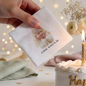 May include: A white square napkin with a printed image of a smiling person wearing a party hat. The text "LOOK WHO'S 30" is printed below the image. The napkin is held near a birthday cake with a lit candle.