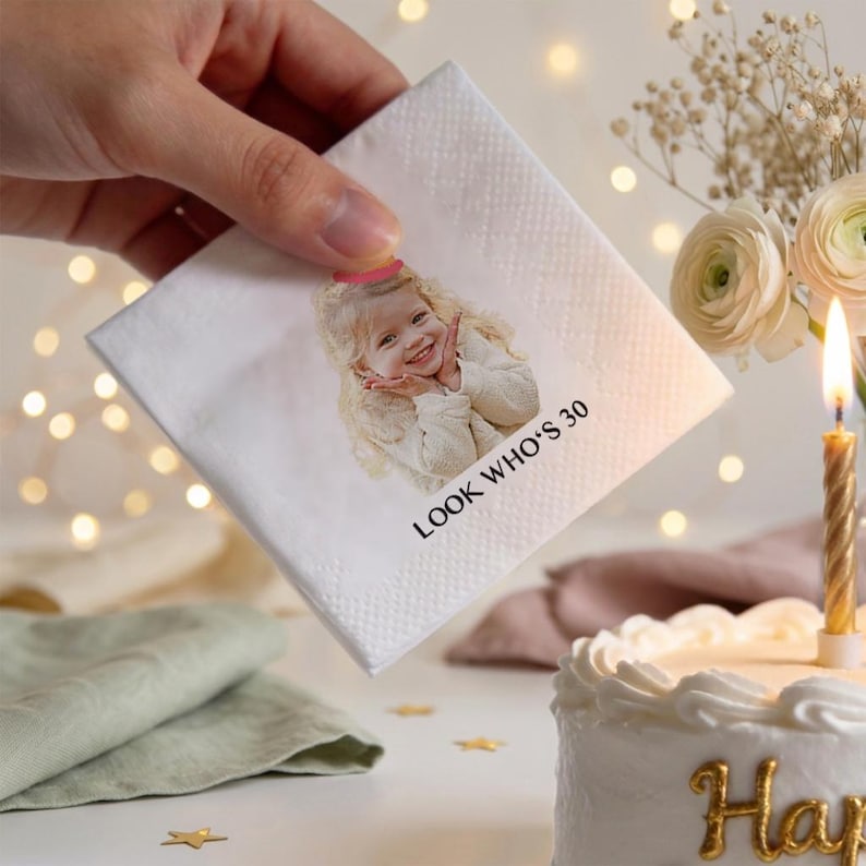 May include: A white square napkin with a printed image of a smiling person wearing a party hat. The text "LOOK WHO'S 30" is printed below the image. The napkin is held near a birthday cake with a lit candle.