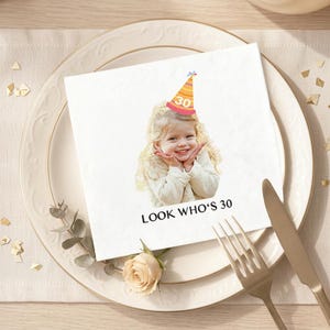 May include: A white paper napkin with a printed image of a smiling child wearing a party hat with the number 30. The text "LOOK WHO'S 30" is printed below the image. The napkin is placed on a white plate with gold trim, next to a fork and knife.