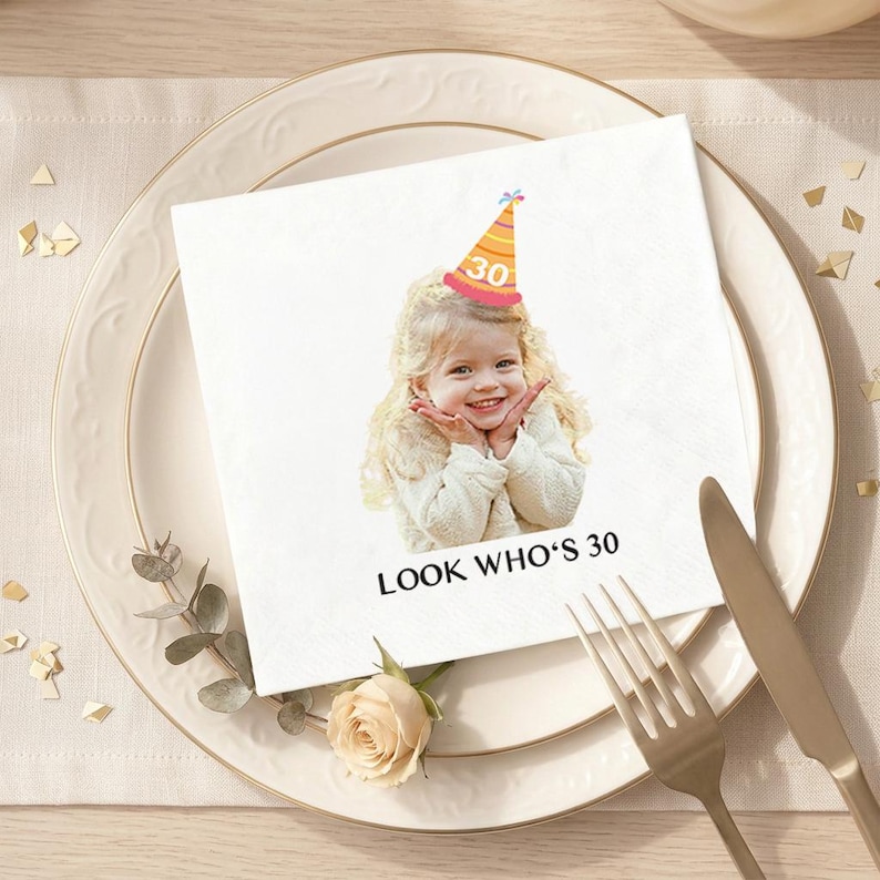 May include: A white paper napkin with a printed image of a smiling child wearing a party hat with the number 30. The text "LOOK WHO'S 30" is printed below the image. The napkin is placed on a white plate with gold trim, next to a fork and knife.