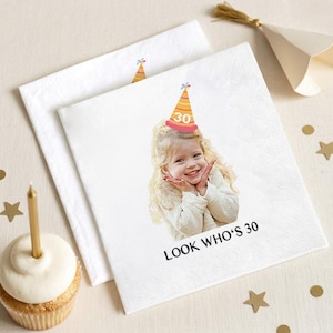 May include: White paper napkins with a printed image of a smiling child wearing a party hat with the number 30. The text "LOOK WHO'S 30" is printed below the image. A cupcake with a gold candle is in the foreground.