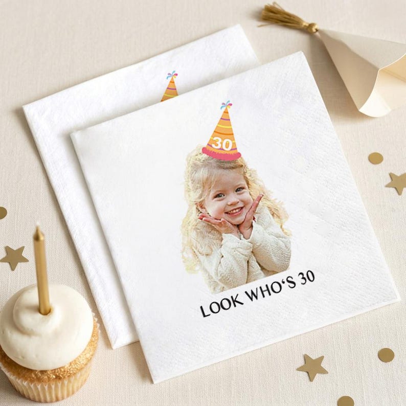 May include: White paper napkins with a printed image of a smiling child wearing a party hat with the number 30. The text "LOOK WHO'S 30" is printed below the image. A cupcake with a gold candle is in the foreground.