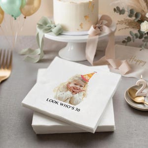 May include: White paper napkins with a printed image of a smiling child wearing a party hat that says "30". The text "LOOK WHO'S 30" is printed below the image. A white cake on a stand and balloons are in the background.