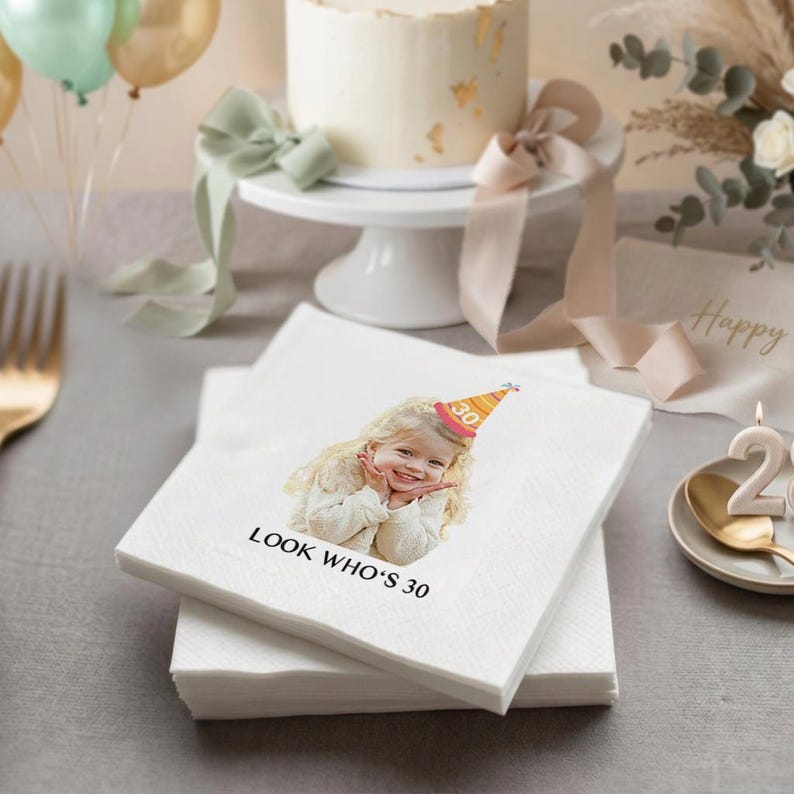May include: White paper napkins with a printed image of a smiling child wearing a party hat that says "30". The text "LOOK WHO'S 30" is printed below the image. A white cake on a stand and balloons are in the background.