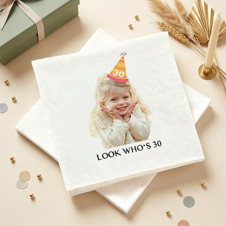 May include: White square paper napkins with a printed image of a smiling child wearing a party hat with the number 30. The text "LOOK WHO'S 30" is printed below the image. The napkins are stacked on top of each other.