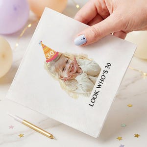 May include: White paper napkin with a photo of a smiling child wearing a party hat with the number 30. The text "LOOK WHO'S 30" is printed on the napkin. A gold candle and star-shaped confetti are also visible.