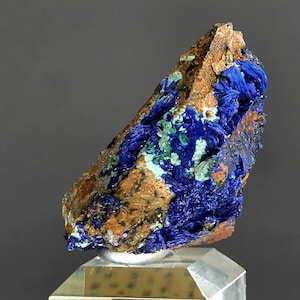 May include: A striking mineral specimen featuring a mix of colors. The piece showcases a rough, brown matrix with vibrant blue and green crystalline formations. The mineral is displayed on a clear, rectangular stand, highlighting its natural beauty.