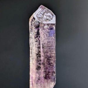 May include: A translucent, elongated crystal with a pointed tip. The crystal displays a gradient of colors, transitioning from a pale lavender at the base to a deeper purple near the center and top. The surface has a textured appearance.
