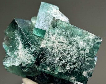 Könnte beinhalten: Nahaufnahme einer Fluorit-Mineralprobe. Der Fluorit weist eine Ansammlung von durchscheinenden, kubischen Kristallen in Grün- und Türkistönen auf. Die Kristalle haben ein mattiertes Aussehen mit sichtbaren inneren Strukturen.