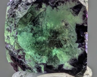 Könnte beinhalten: Ein großer, grüner Fluoritkristall mit violetten und schwarzen Akzenten. Der Kristall ist würfelförmig und hat eine glatte, polierte Oberfläche.