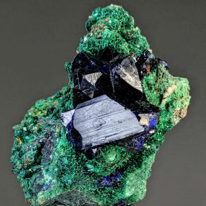 May include: A close-up of a mineral specimen featuring a cluster of dark blue, crystalline structures set against a backdrop of vibrant green, needle-like formations. The mineral has a metallic sheen and is set against a dark gray background.