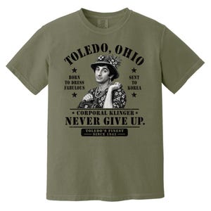 May include: An olive green short-sleeve t-shirt featuring a black graphic print of Corporal Klinger. The text reads: TOLEDO, OHIO, BORN TO DRESS FABULOUS, SENT TO KOREA, CORPORAL KLINGER NEVER GIVE UP, and TOLEDO'S FINEST SINCE 1943 with star accents.