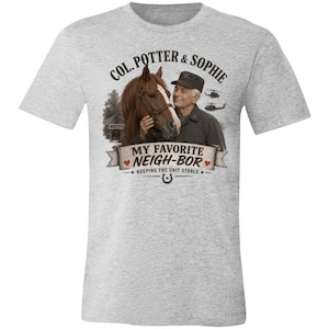 May include: A heather gray t-shirt featuring a graphic of a person in a dark uniform standing next to a brown horse. The text reads COL. POTTER & SOPHIE, MY FAVORITE NEIGH-BOR, and KEEPING THE UNIT STABLE, with small helicopters and a 4077 sign.