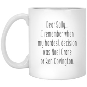 May include: A white ceramic mug featuring black printed text that reads: Dear Sally... I remember when my hardest decision was Noel Crane or Ben Covington. The mug has a standard curved handle on the left side and a smooth, cylindrical body.