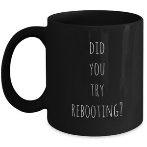 May include: Black ceramic coffee mug with a white handle. The mug features the text "DID YOU TRY REBOOTING?" in a vertical stack, using a simple sans-serif font. The mug is set against a white background.
