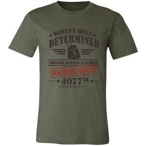 May include: An olive green short-sleeve t-shirt featuring a dark graphic print that reads "WORLD'S MOST DETERMINED," "CORPORAL MAXWELL Q. KLINGER," "DISCHARGE DENIED," and "4077TH M*A*S*H," along with an illustration of a dog tag labeled "SECTION 8."
