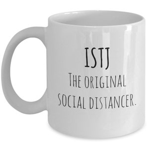 Puede incluir: Taza de cerámica blanca con asa curvada. La taza presenta el texto negro "ISTJ The ORIGINAL SOCIAL DISTANCER" centrado en la parte delantera. La taza es de tamaño estándar para café o té.